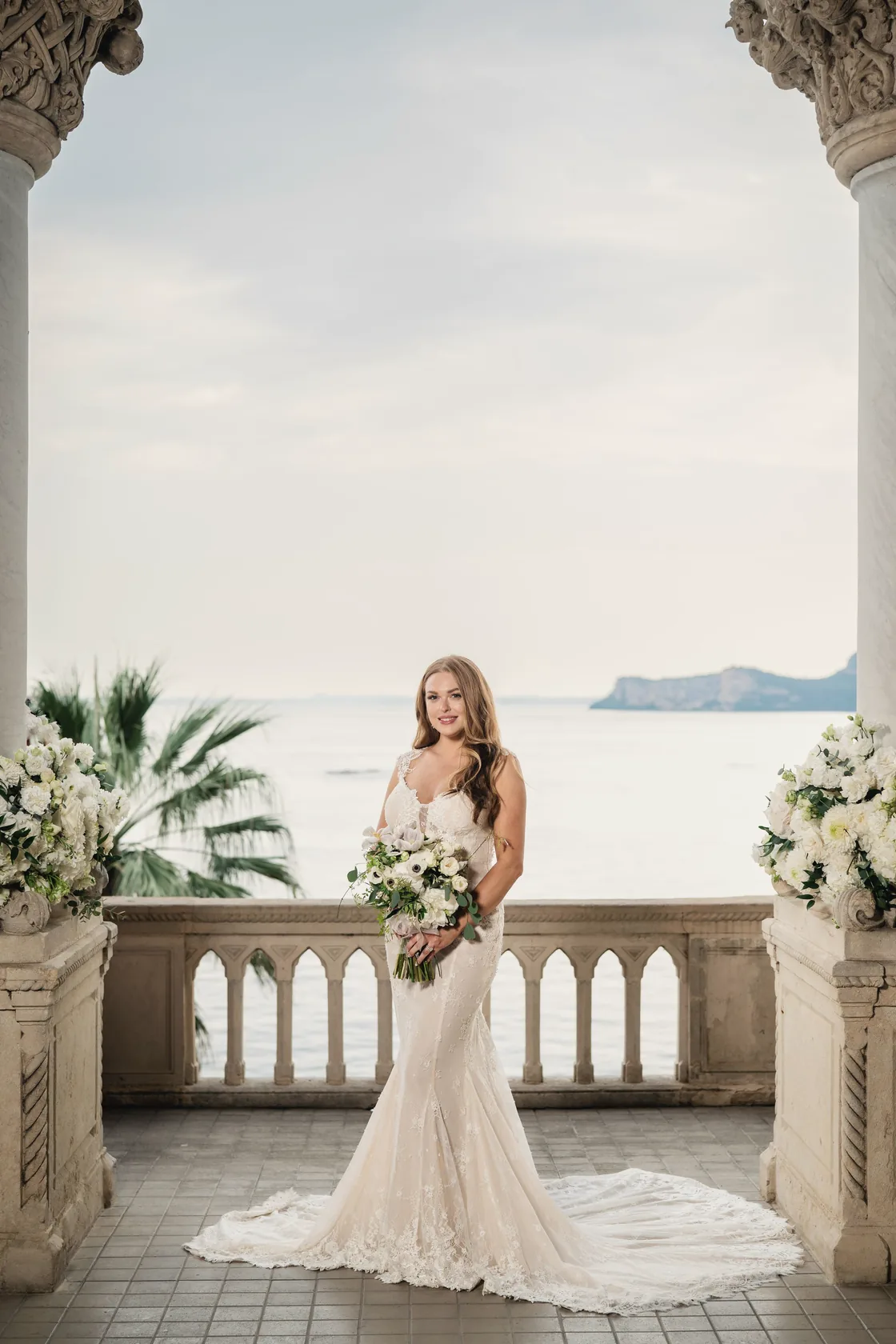 A bride stands alone beneath an arch that frames the lake and distant hills