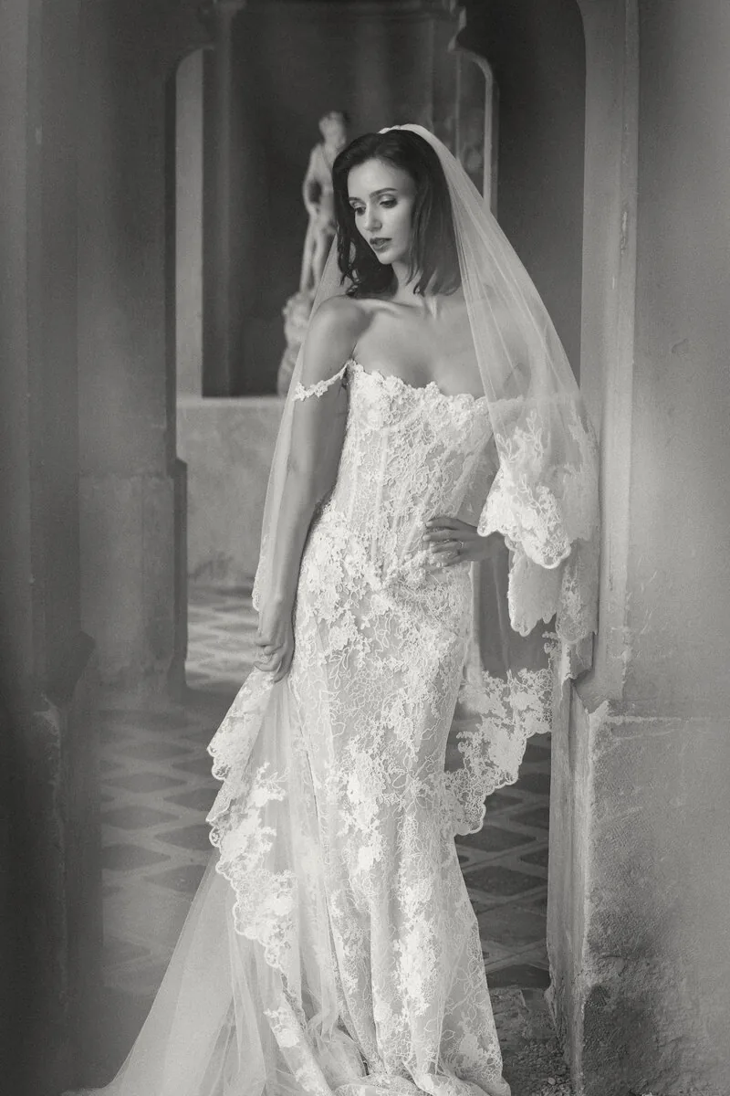 Black and white portrait of a bride in a lace gown and long veil in a stone colonnade