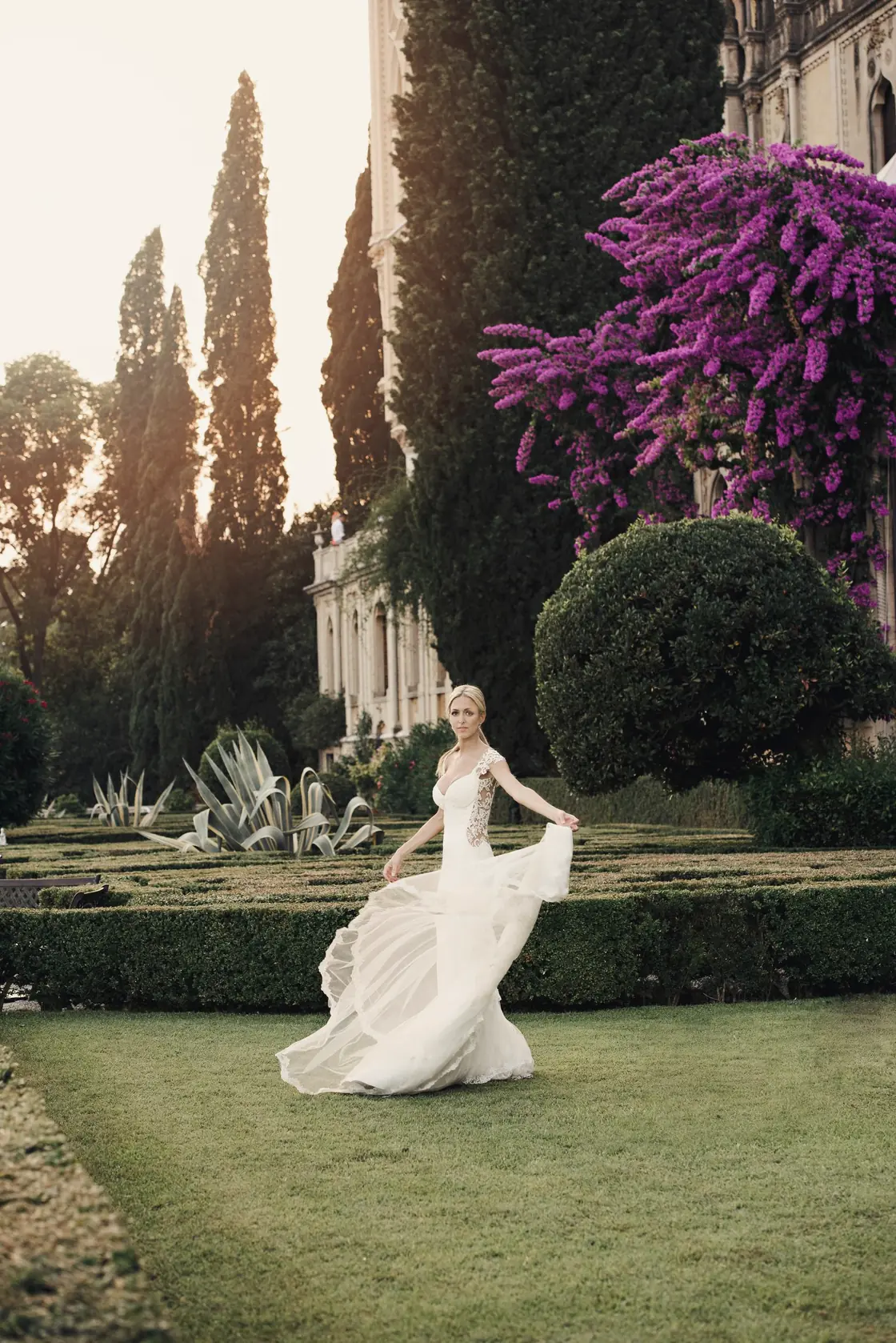 A bride lifts her skirt as she walks through a formal garden past pink bougainvillea