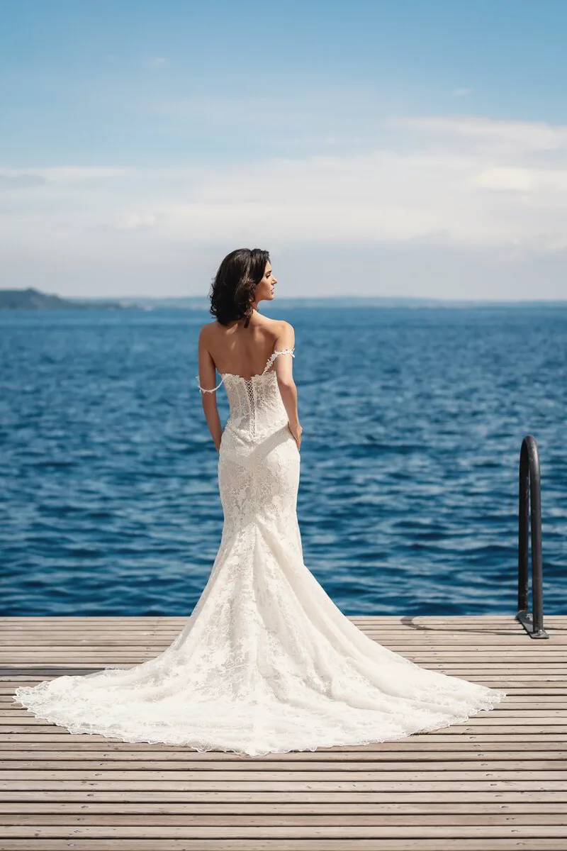 A bride in a lace gown with a long train stands on a wooden jetty, looking out over a deep blue lake