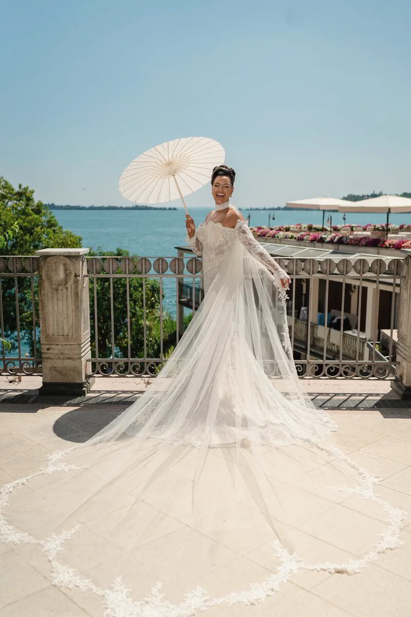 A bride with a white parasol and a long cathedral veil on a lakeside terrace