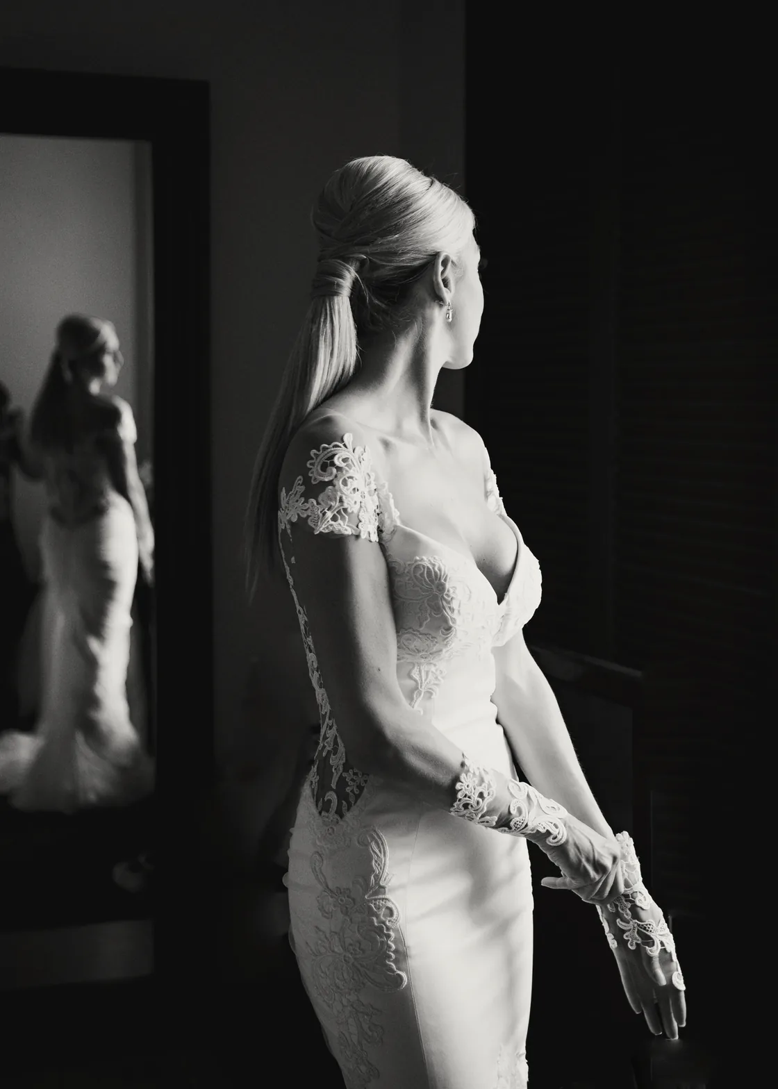 A bride in a lace gown turns towards the window of a darkened room