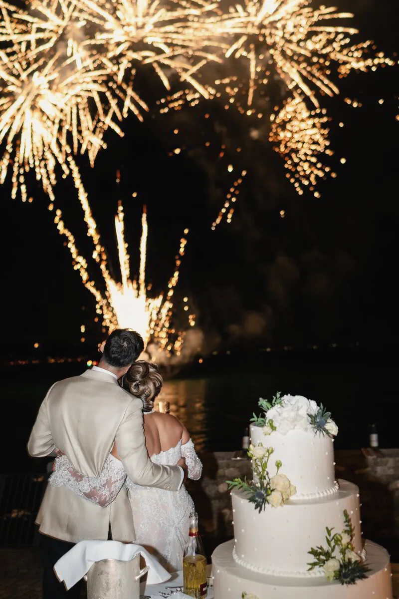 Fireworks burst over a couple cutting a tiered white cake at night
