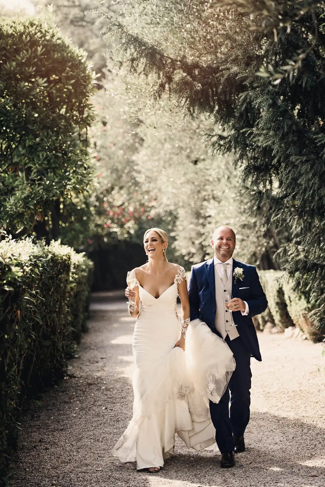 A bride and groom walk hand in hand down a shaded garden path