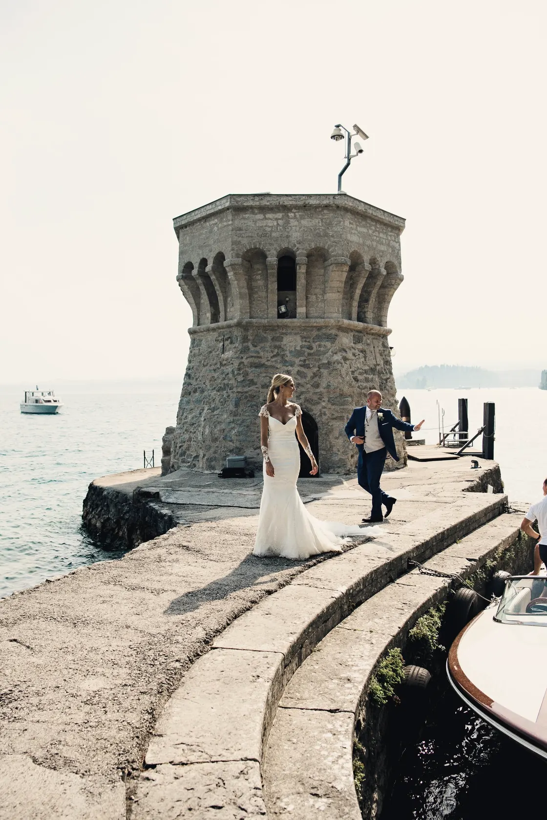 A couple walks along a curved stone pier beside a round harbour tower
