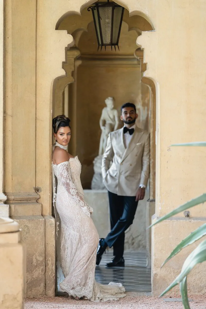 A couple stands in a sunlit stone archway, the bride in a sheer embroidered gown