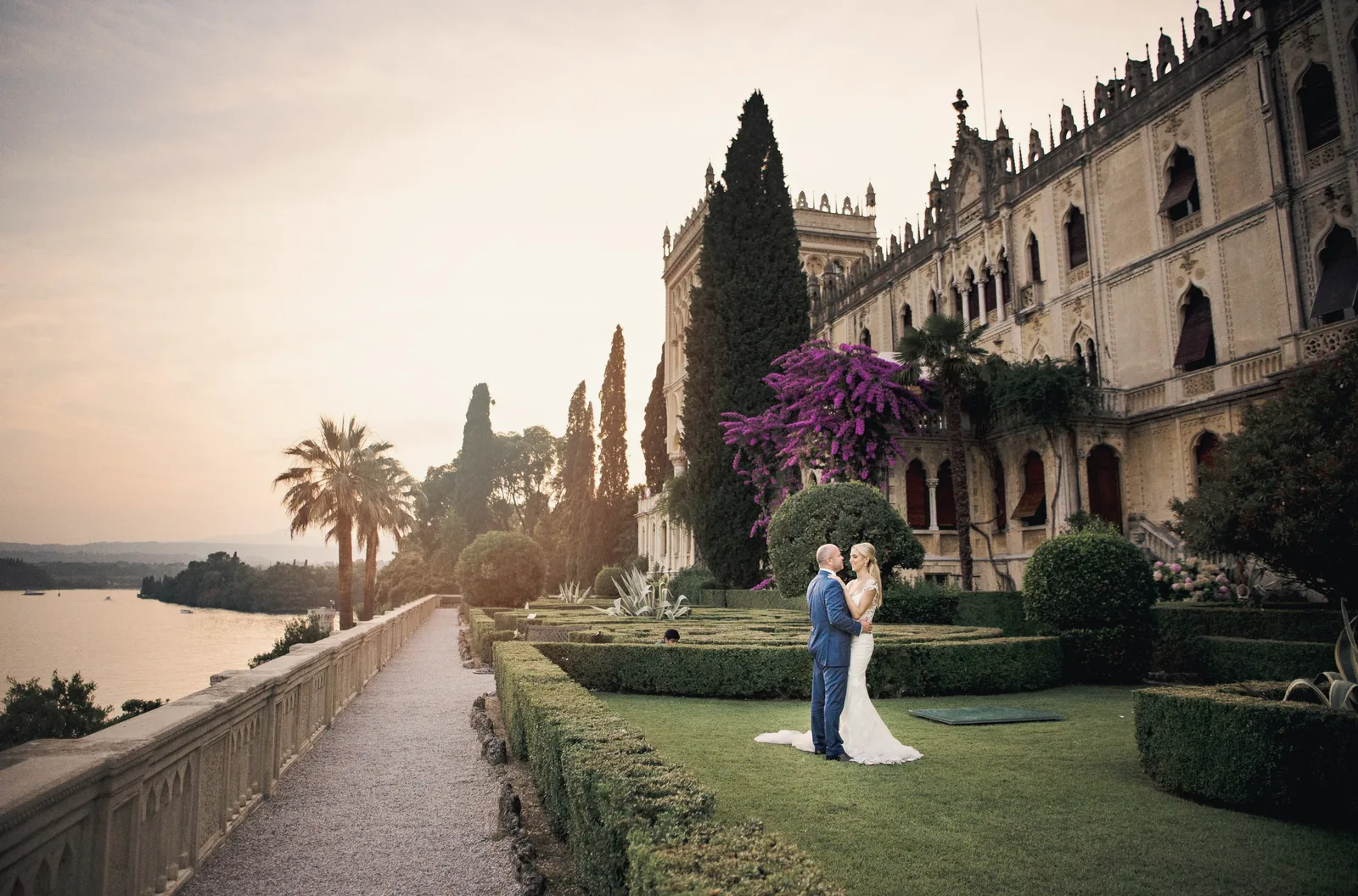 A couple stands in a formal lakeside garden as the sun sets behind cypress trees and a villa