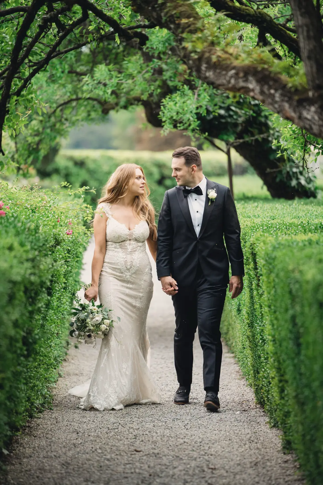 A couple walks between clipped hedges beneath an arch of trees