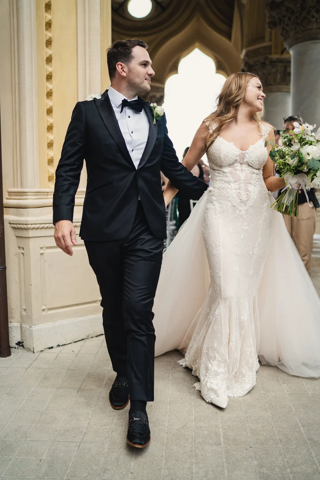 A newly married couple walks out of a Gothic arcade, the bride holding her bouquet