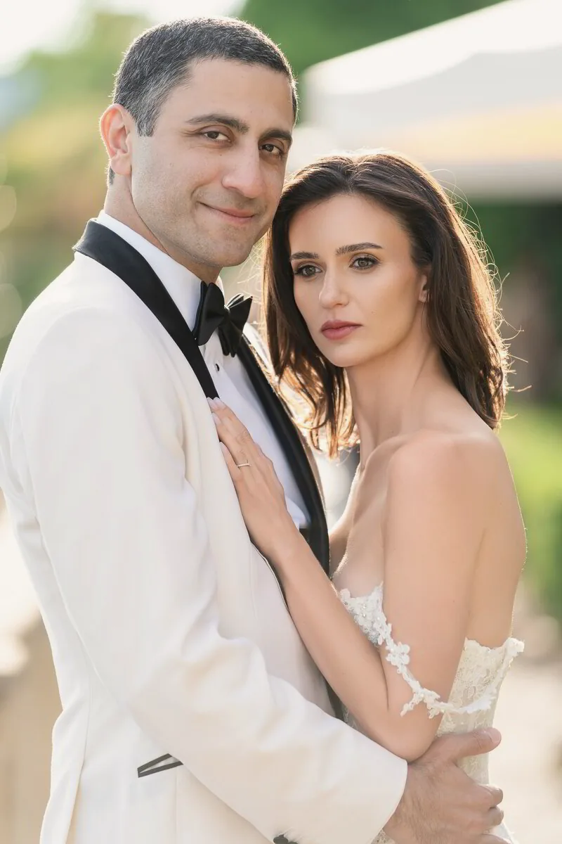 A groom in a white dinner jacket holds the bride close as they both look into the camera