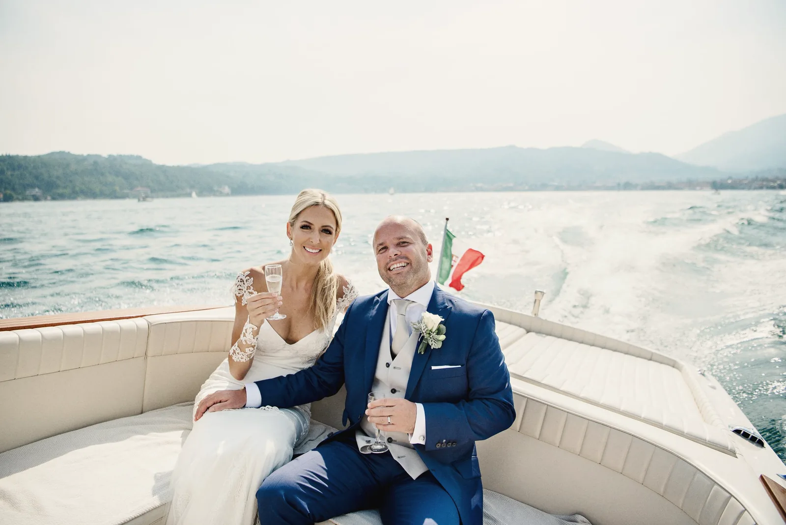 A bride and groom laugh in the back of a wooden motorboat crossing the lake
