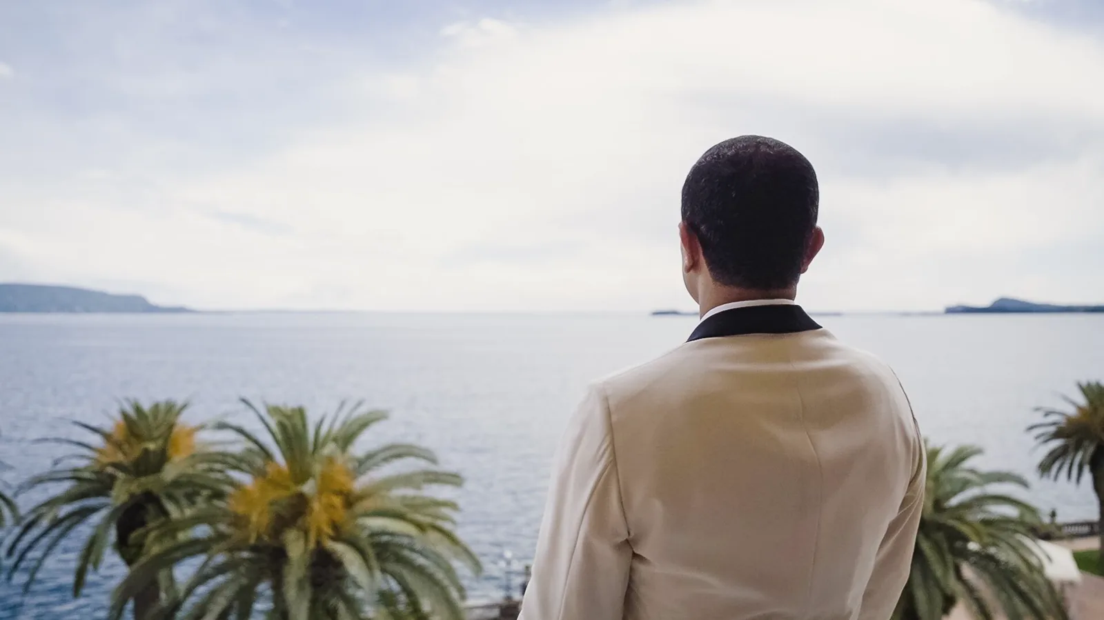 A groom in an ivory jacket looks out over the lake between palm trees