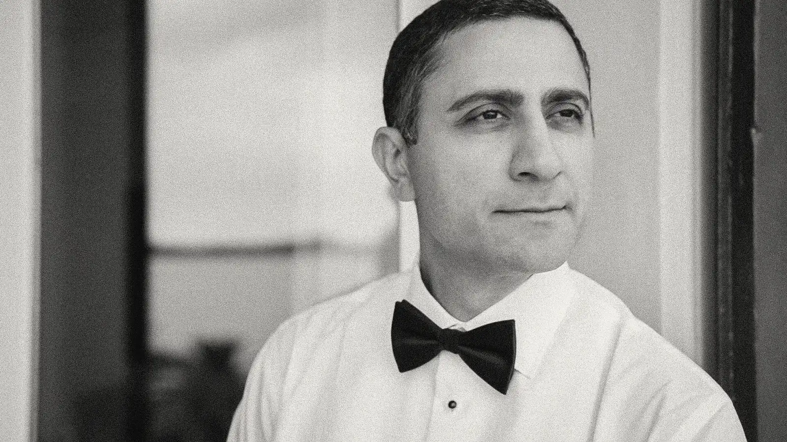 Black and white portrait of a groom in a bow tie beside a window