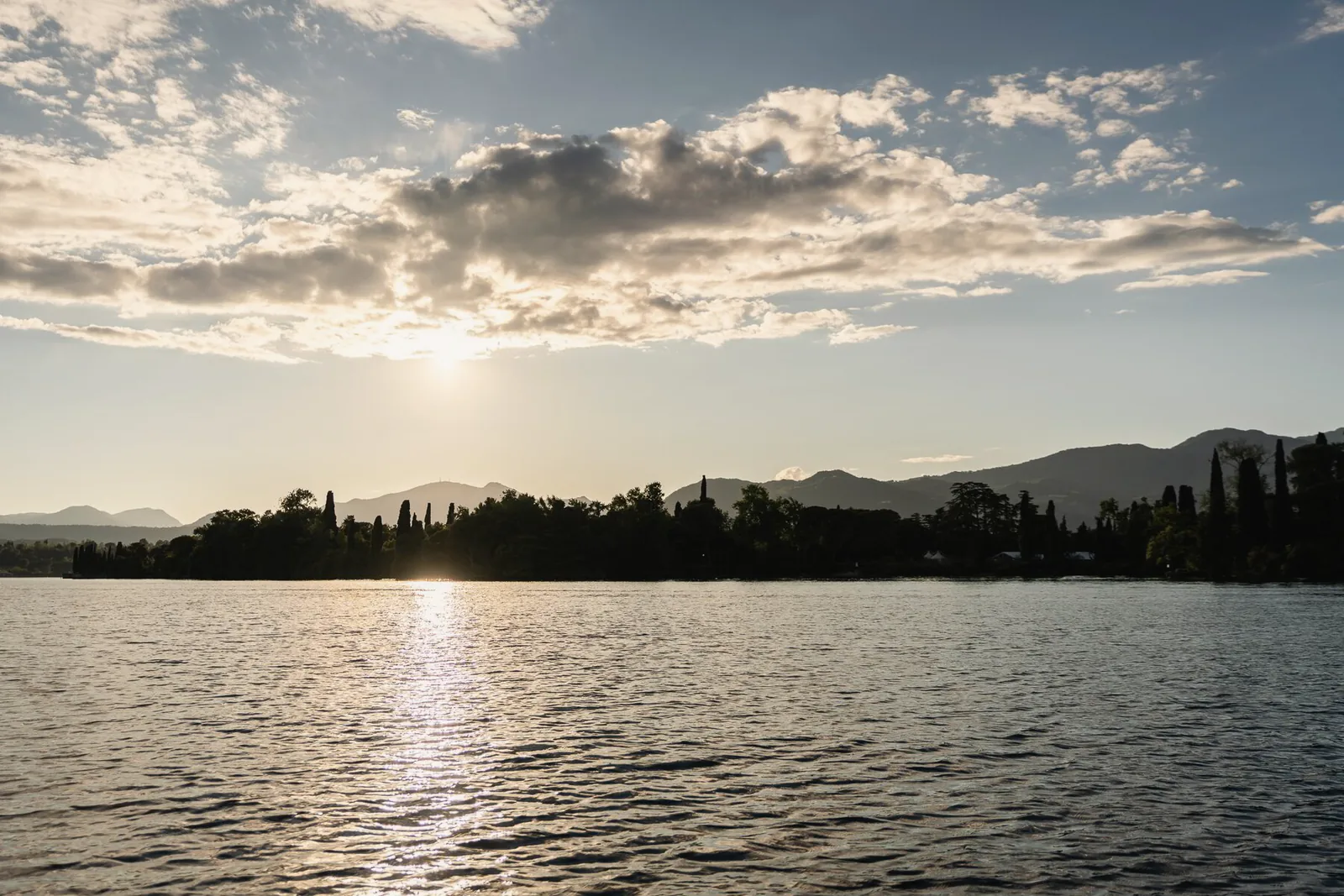 The sun sets over a still lake with low hills on the horizon