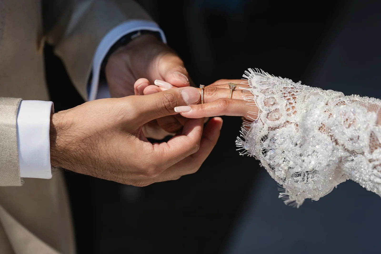 A groom slides a ring onto the bride's finger, her sleeve covered in lace